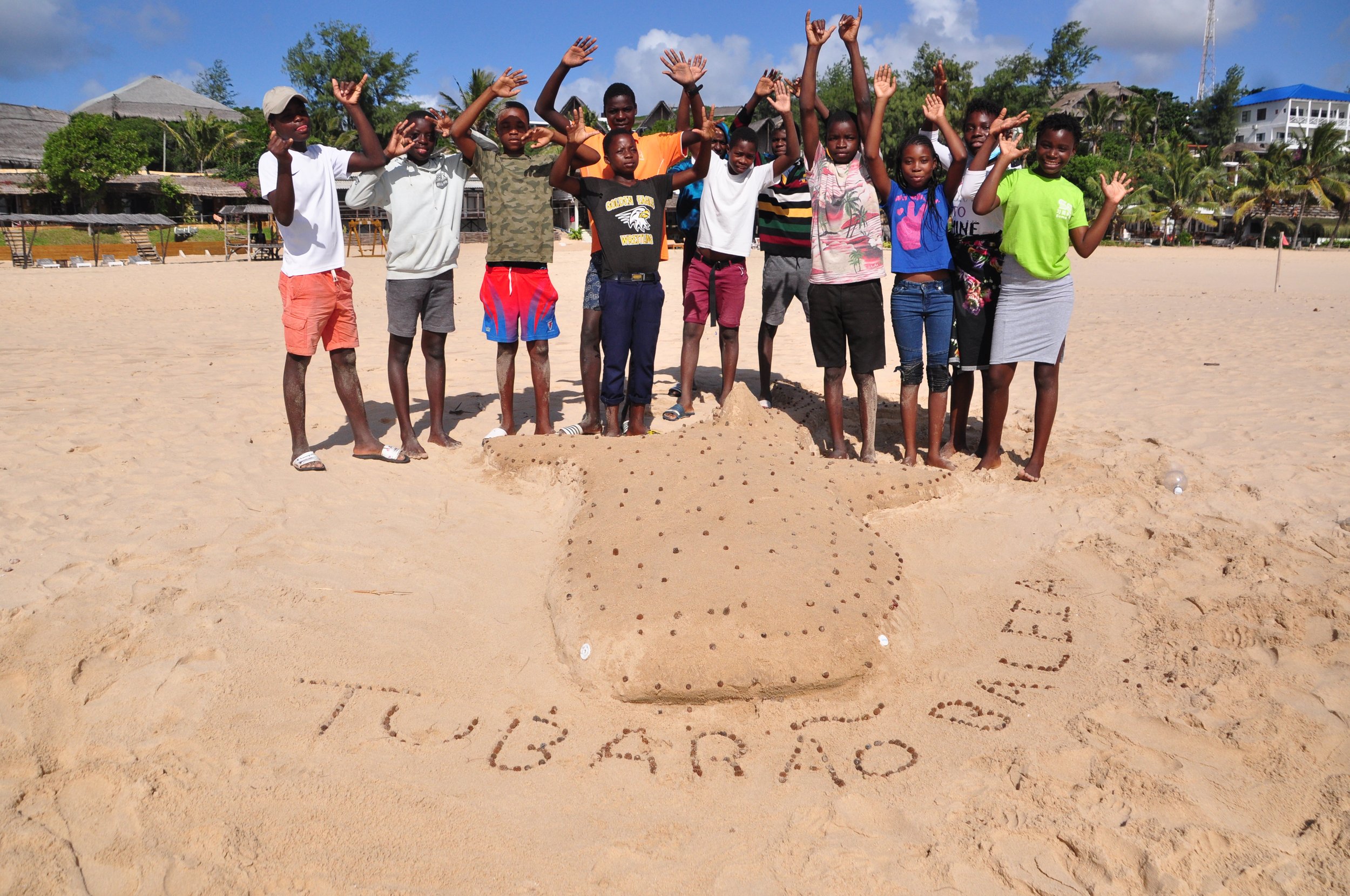 Children cheer behind a whale shark they sculpted in the sand, with "Tubarão Baleia" written beside it